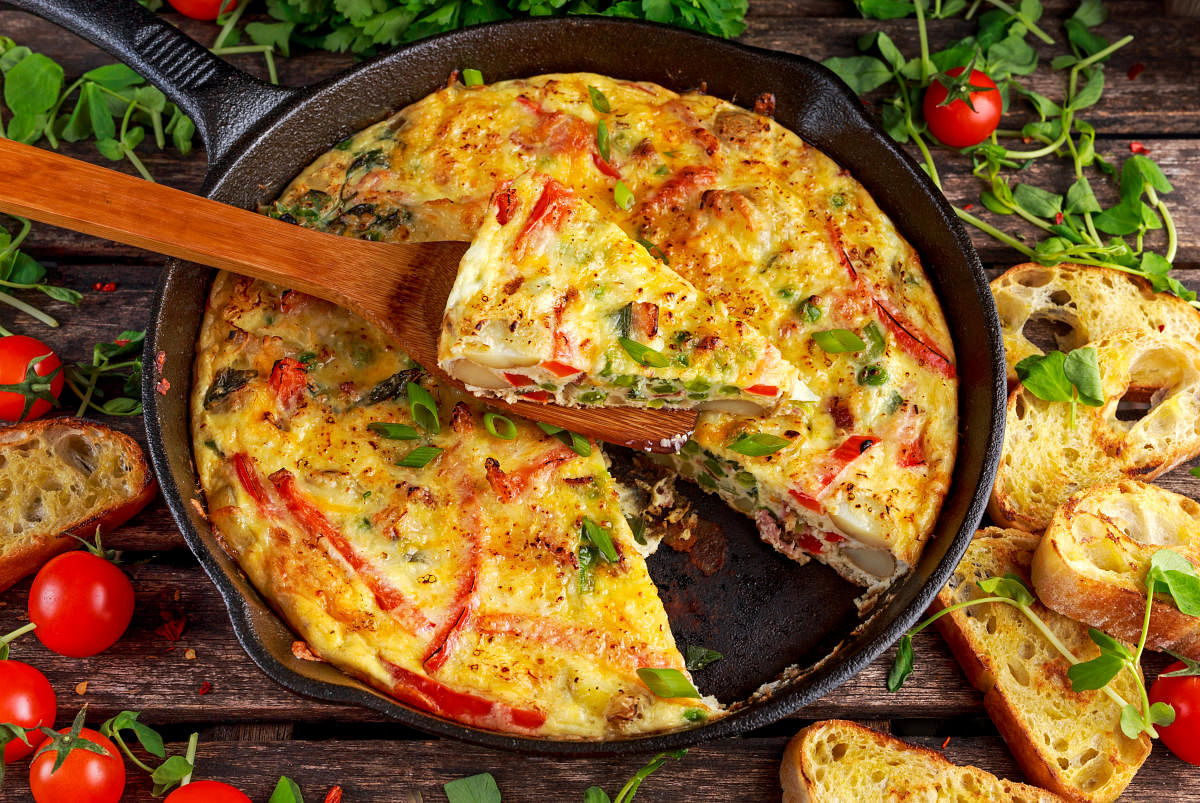 Frittata made of eggs, potato, bacon, paprika, parsley, green peas, onion, cheese in iron pan on wooden table